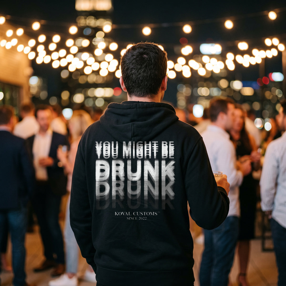 Man at a rooftop party in a black hoodie with a motion-blur You Might Be Drunk print across the back