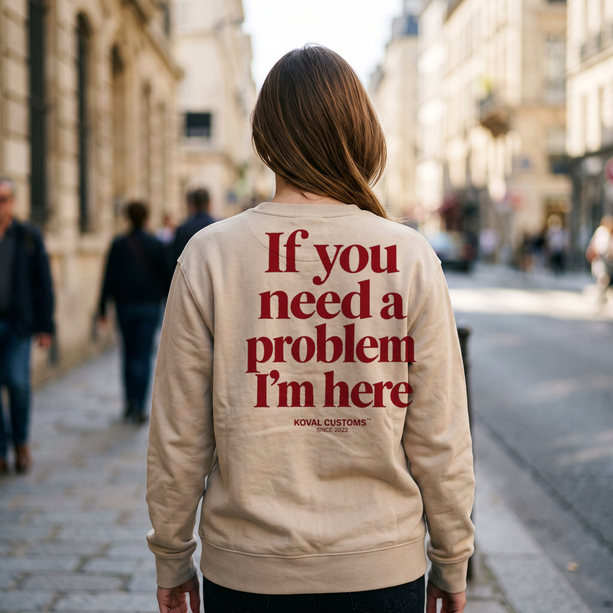 Woman walking a sunlit city street in a sand crewneck sweatshirt with If You Need A Problem I'm Here printed across the back