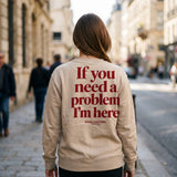 Woman walking a sunlit city street in a sand crewneck sweatshirt with If You Need A Problem I'm Here printed across the back