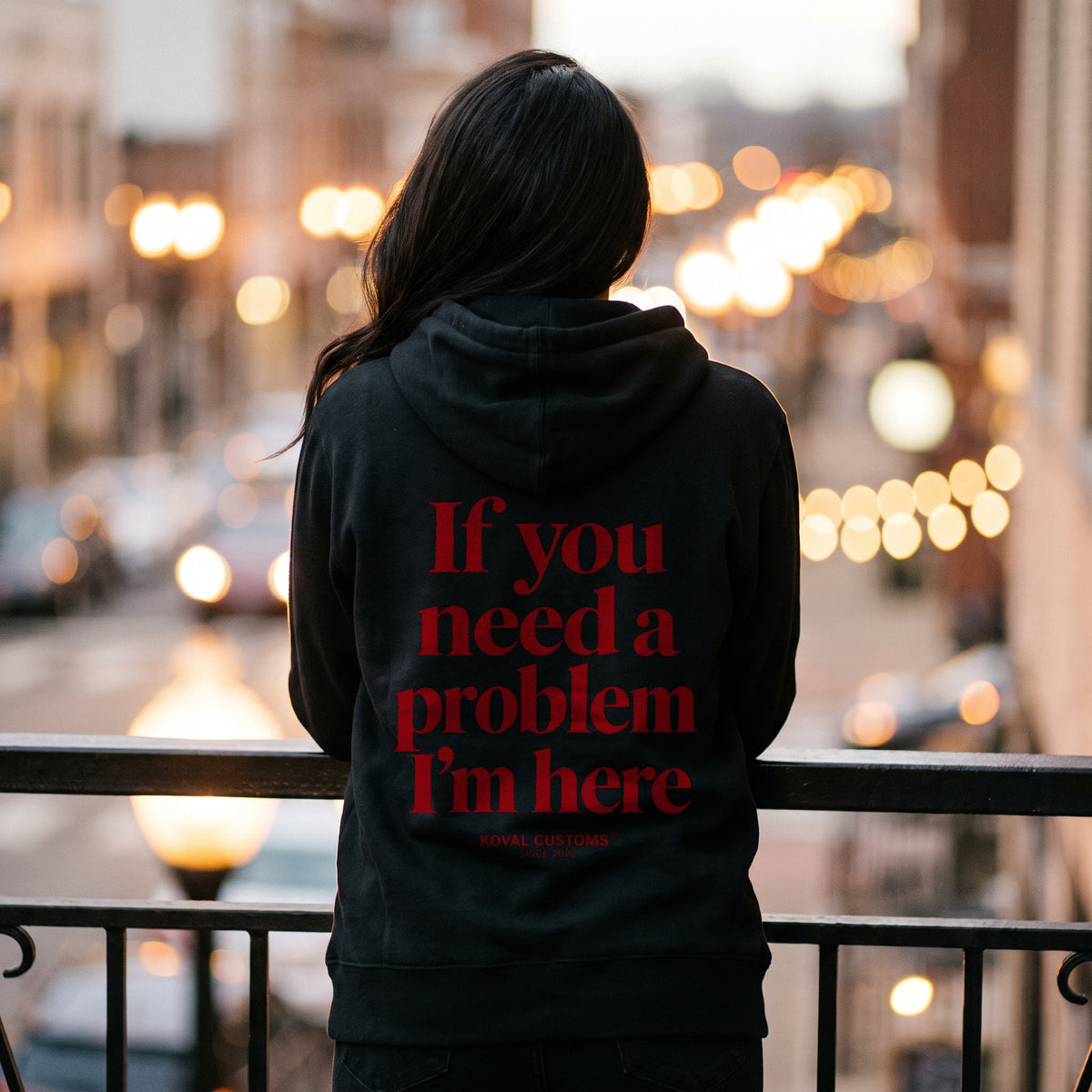 Woman on a balcony in the evening in a black hoodie with If You Need A Problem I'm Here printed across the back