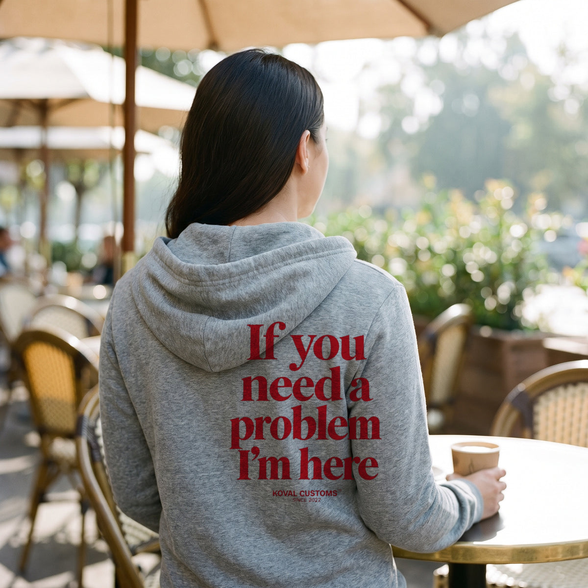 Woman at an outdoor cafe in a sport grey zip hoodie with If You Need A Problem I'm Here printed across the back